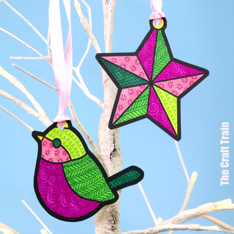 Christmas pattern art paper star and bird decorations hung with pink ribbon on a white branch before a pale blue background. The decorations are coloured in shades of pink and green, then decorated with detailed doodle art patterns.