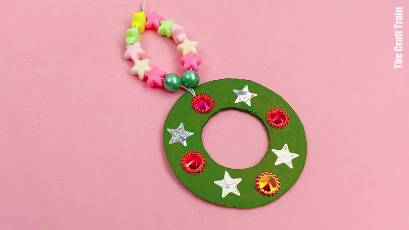 Finished cardboard Christmas decoration craft, photographed on a pink background. Recycled cardboard has been cut into the shape of a wreath, painted green, then decorated with rhinestones and sequins. The string to hang it has been threaded with plastic coloured beads