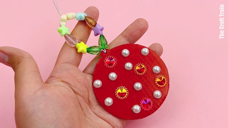Finished cardboard Christmas decoration craft, photographed on a pink background. Recycled cardboard has been cut into the shape of a round bauble, painted red, then decorated with rhinestones and sequins. The string to hang it has been threaded with plastic coloured beads