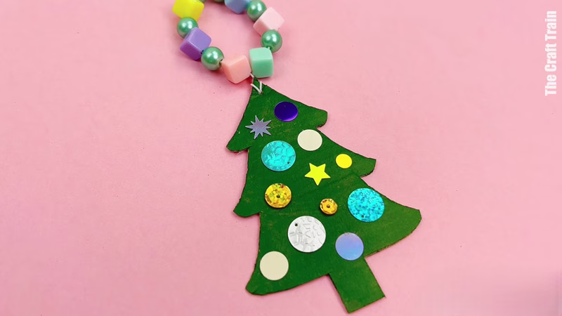 Finished cardboard Christmas decoration craft, photographed on a pink background. Recycled cardboard has been cut into the shape of a tree, painted green, then decorated with rhinestones and sequins. The string to hang it has been threaded with plastic coloured beads