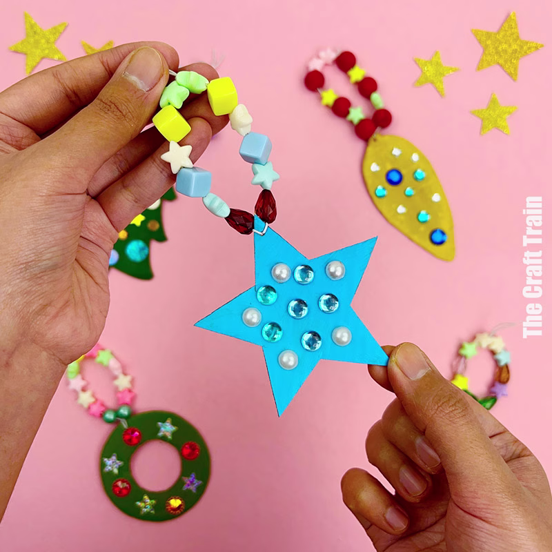 Recycle cardboard Christmas ornaments. A handmade blue painted cardboard star shape with beaded string is being held, surrounded by other handmade Christmas shape ornaments.