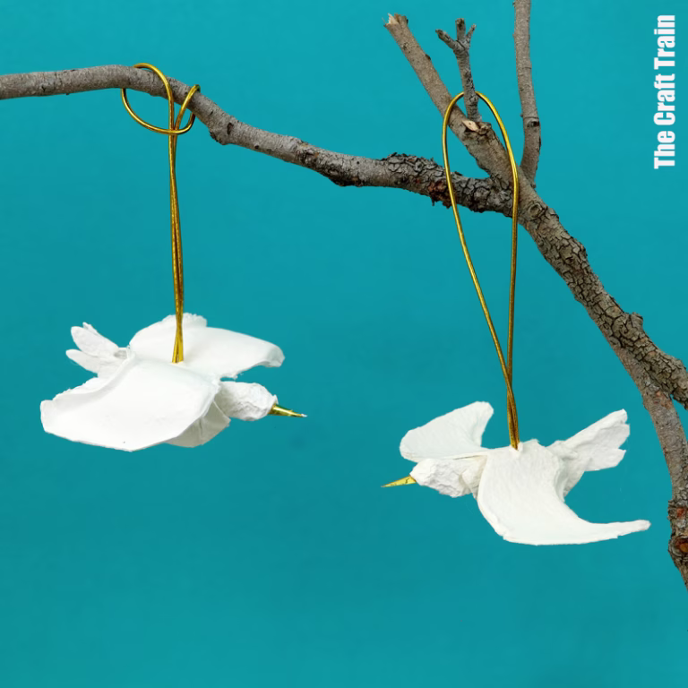Handmade egg carton peace dove ornaments. There are two doves hanging from a branch by a gold thread, photographed against a teal backdrop